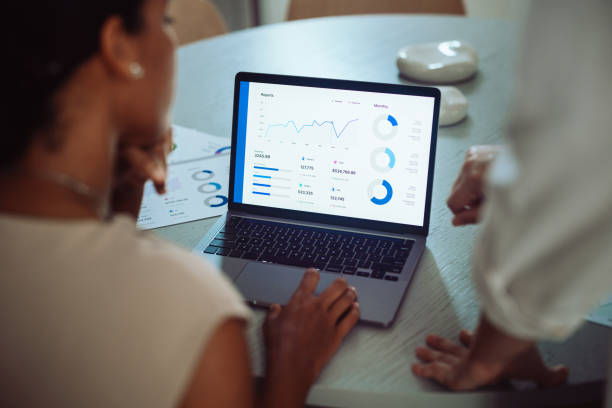 Two business professionals analyzing sales data and financial charts on a laptop during a meeting in a modern office setting.